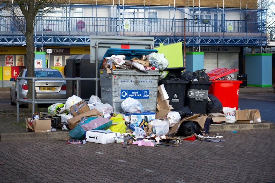 A collection of discarded household waste, including a small evergreen tree, a cardboard box with crumpled paper, a black recycling bag, and a large white parcel box with a blue recycling symbol, plac