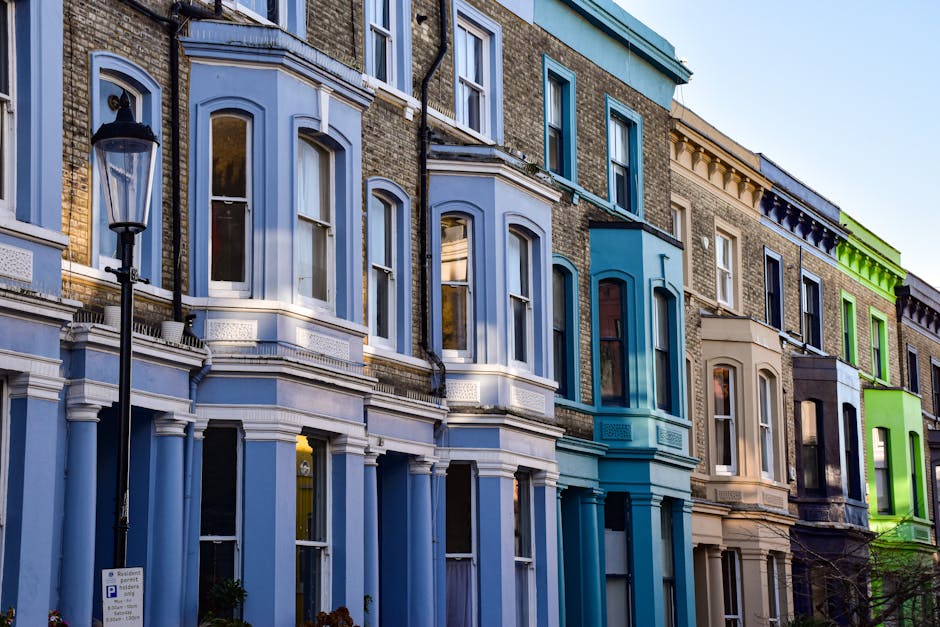 A row of Victorian-style terraced houses with three to four stories, featuring a combination of painted and exposed brick façades. The buildings display a variety of pastel shades, including white, p